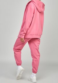Roze hoodie en bijpassende joggingbroek van zacht materiaal, met een relaxte pasvorm, geribbelde boorden en een capuchon. Gedragen met witte sneakers.