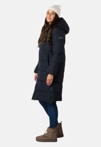 Lang marineblå pufferjakke med hætte, med quiltet design, sidelommer og to forlommer. Parret med beige vinterstøvler og jeans.