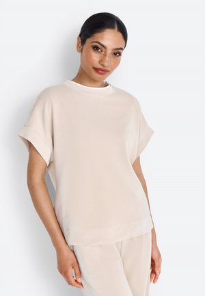 Woman with dark hair in a low bun wearing a beige short-sleeve top and matching pants, standing against a plain light background.