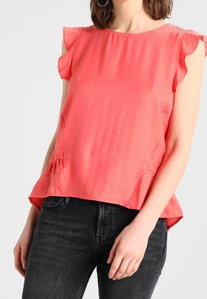 Femme portant un chemisier sans manches corail avec des manches courtes à volants, associé à un jean noir taille haute, debout devant un fond uni.
