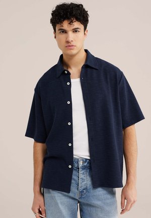 Jonge man met kort, krullend haar, gekleed in een marineblauw kortärmelig overhemd met knopen, gedragen over een wit tanktop en lichtblauwe spijkerbroek, tegen een beige achtergrond.