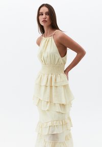 Žluté maxi šaty s vrstveným střihem, pevným smockovaným pasem, halter krkem a volánkovými vrstvami, vyrobené z lehkého materiálu.