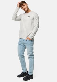 Lys grå langærmet T-shirt med et lille sort logo på brystet, parret med lyseblå denimjeans og sorte sneakers.
