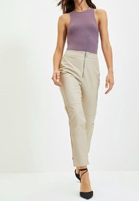 Vrouw die wandelt in beige high-waist broek met een rits aan de voorkant, mouwloos paarse top en zwarte puntige hakken met enkelbandjes.
