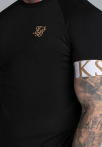 Melna īsām piedurknēm T-kreklu ar zeltītu izšuvumu logo kreisajā krūtī un kontrastējošu baltu joslu ar zelta burtiem uz piedurknes.