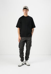 T-shirt noir à manches courtes, pantalon cargo gris avec poches, baskets blanches avec rayures noires, et casquette gris clair. Fond uni.