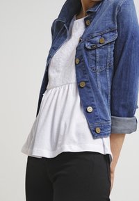 Giacca di jeans blu con bottoni dorati, due tasche frontali e maniche arrotolate, sopra una blusa bianca a strati con una texture in pizzo. Pantaloni neri.