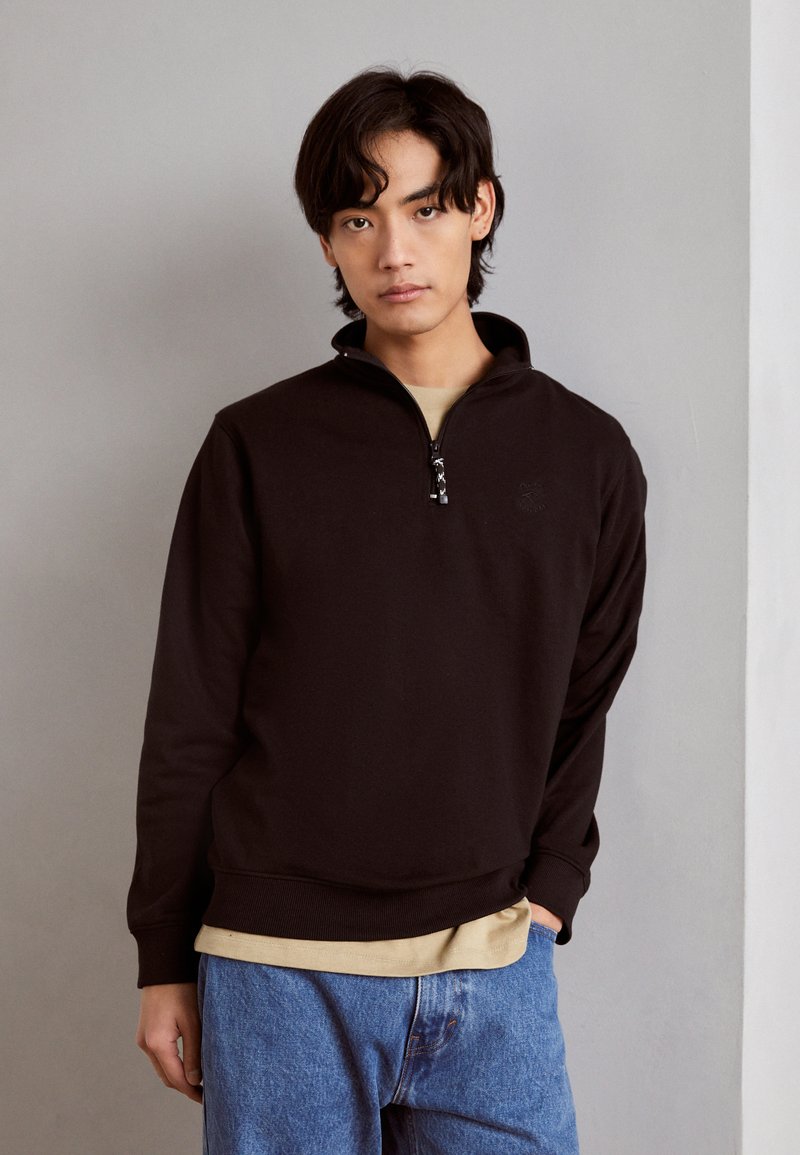Sort halv-zip trøje lavet af blødt stof, med ribkant ved kanten og manchetterne, lagdelt over en lys beige skjorte, parret med blå jeans.