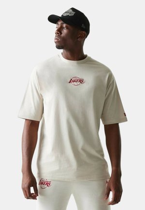 Uomo che indossa una maglietta beige dei Los Angeles Lakers e pantaloni, abbinati a un cappellino da baseball nero dei Lakers, che guarda verso sinistra.