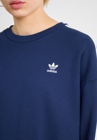 Marinblå sweatshirt med rund halsringning. Har vit broderad Adidas-logga på vänster sida. Mjukt tyg och avslappnad passform.