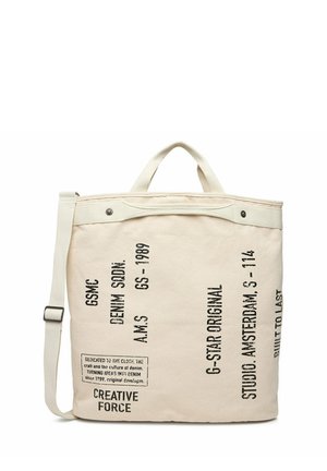 Cream canvas tote bag with black printed text, dual handles, and an adjustable shoulder strap, featuring branding and design details.