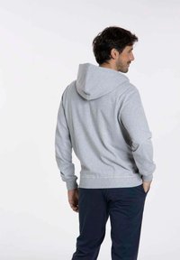 Sweatshirt gris avec capuche, poignets et taille côtelés. Le tissu semble doux et texturé, présenté de dos avec une coupe décontractée.