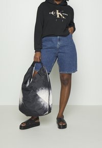 Svart bomullströja med logotyp, blå denimshorts och svarta sandaler. Väskan har ett tie-dye-mönster i svart och vitt.