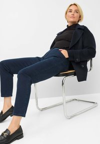 Navy wollen blend jas met brede revers, zwarte coltrui, donkerblauwe jeans en zwarte instappers met een gouden accent, zittend op een chroomstoel.