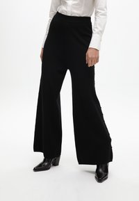 Černé široké kalhoty vyrobené z měkké tkaniny, s vysokým pasem a ozdobnými knoflíky po straně. Vzájemně sladěné s černými botami na podpatku.