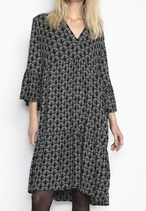 Frau trägt ein schwarz-weiß gemustertes Midikleid mit 3/4-Ärmeln und V-Ausschnitt vor einem einfachen weißen Hintergrund.