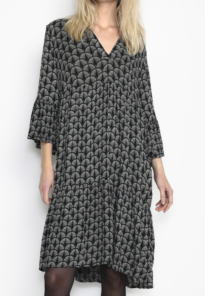 Femme portant une robe midi à motifs noir et blanc avec manches trois-quarts et col en V, sur un fond blanc uni.