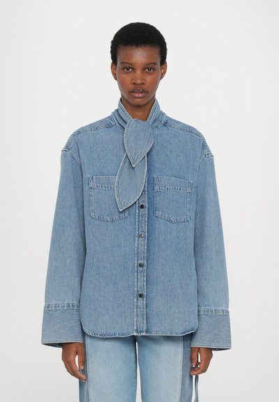 Camisa de denim azul claro con un gran cuello tipo solapa, dos bolsillos en el pecho y cierre frontal de botones. Mangas largas con puños estructurados.