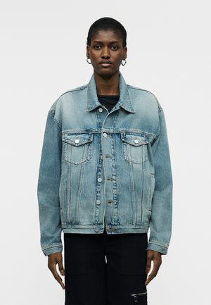 Γυναίκα που φοράει ανοιχτό γαλάζιο φαρδύ τζιν μπουφάν με μπροστινές τσέπες και μαύρο παντελόνι, στέκεται μπροστά σε απλό φωτεινό φόντο.