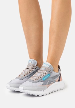 Personne portant des baskets Reebok Classic grises et rose clair avec des étiquettes de logo bleues, debout sur une surface blanche.