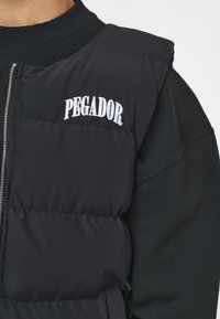 Svart vadderad väst med dragkedja, prydd med "PEGADOR" broderat i vitt. Tyget ser slätt ut med en texturerad, quiltad design.