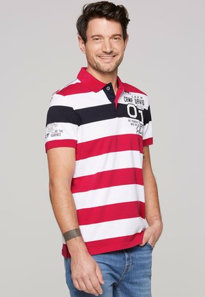 Polo tričko s červenými a bielymi pruhmi, s námořníckou modrou akcentou a golierom. Obsahuje logo na ľavej hrudi a krátke rukávy.