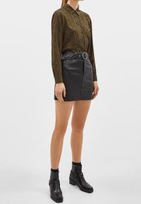 Košilová blůza s dlouhým rukávem v zelené a hnědé zvířecí potisku, spárovaná s černou sukní z faux kůže se zavazovacím pasem. Černé kotníkové boty.