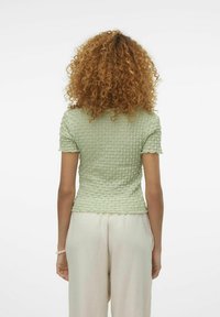 Grön texturerad kortärmad topp med en figurnära silhuett, som har scallopkanter och en krinklad design, tillsammans med ljusa byxor.