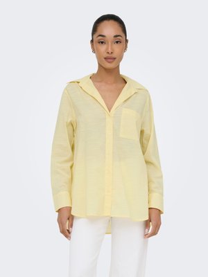 Femme aux cheveux attachés portant une chemise ample boutonnée jaune pâle avec une poche poitrine et un pantalon blanc, debout devant un fond uni.
