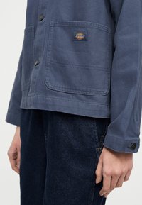 Κοντινό πλάνο ενός ατόμου που φοράει μπλε μπουφάν Dickies με κουμπιά και μπροστινές τσέπες και σκούρο μπλε τζιν, με το χέρι να ακουμπά στο πλευρό του.