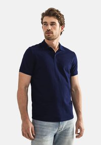 Marineblauw poloshirt van katoen met korte mouwen, drie knopen aan de voorkant en een subtiele merknaam op de linkerborst. Draagt samen met lichte jeans.