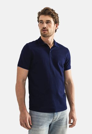 Navy blue polo shirt made of cotton with short sleeves, three-button placket, and subtle branding on the left chest. Worn with light jeans.