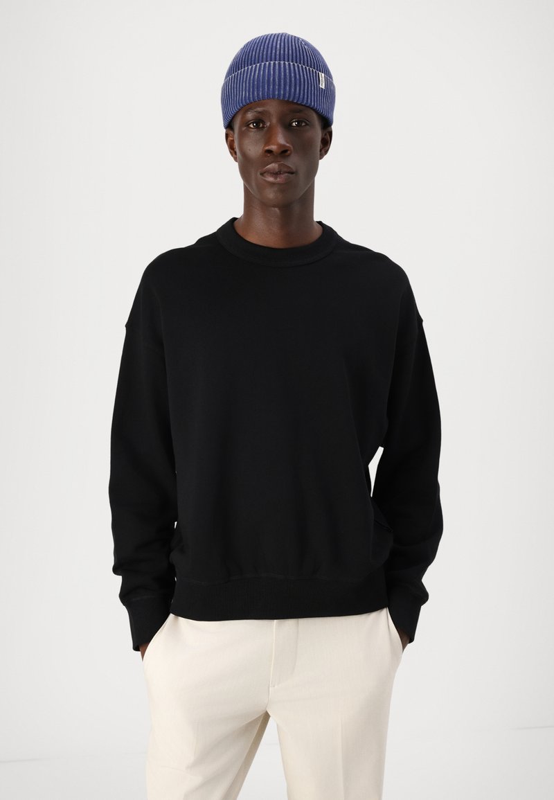 Czarna bluza z okrągłym dekoltem i ściągaczami, w komplecie z jasnobeżowymi spodniami. Model nosi niebieską czapkę z ściągaczem. Gładkie tło.