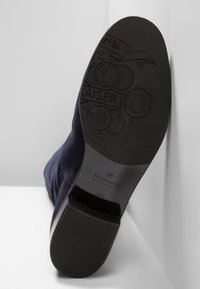 Stivale alla caviglia in suede blu navy con suola piatta in gomma nera. La suola presenta un marchio in rilievo e dettagli decorativi a forma di moneta.