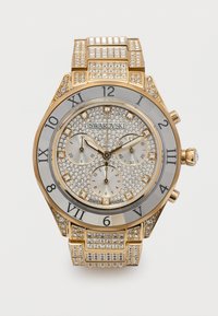 Ρολόι Swarovski σε χρυσό τόνο με καντράν και μπρασελέ επενδυμένα με κρύσταλλα, λατινικούς αριθμούς, τρεις υπογράμματα και σήμανση "κατασκευάστηκε στην Ελβετία" σε λευκό φόντο.