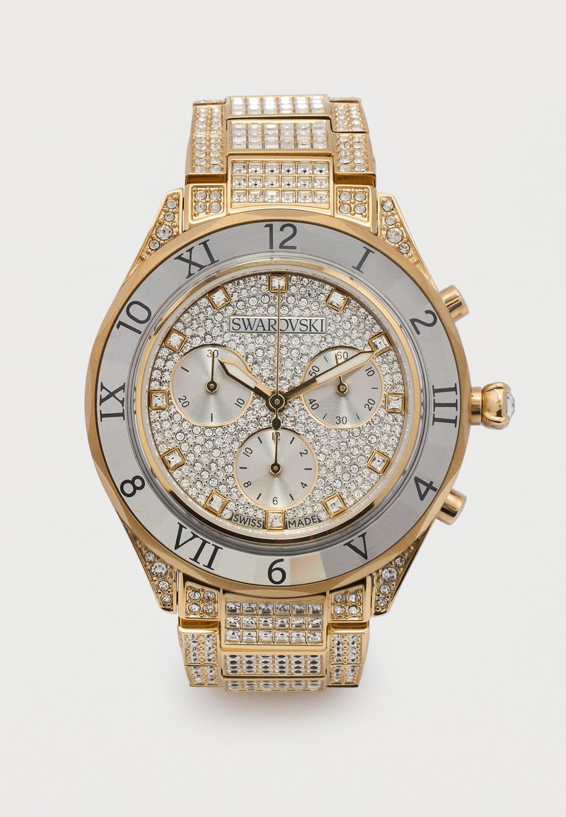 Ρολόι Swarovski σε χρυσό τόνο με καντράν και μπρασελέ επενδυμένα με κρύσταλλα, λατινικούς αριθμούς, τρεις υπογράμματα και σήμανση "κατασκευάστηκε στην Ελβετία" σε λευκό φόντο.