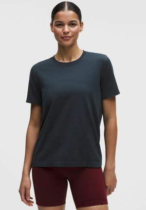 Kobieta z ciemnymi włosami związanymi z tyłu, ubrana w jednolity ciemnoszary krótki rękaw i bordowe spodenki rowerowe, stojąca na neutralnym tle.