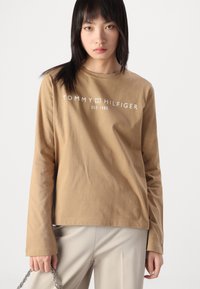 Braunes Langarm-T-Shirt mit einem weißen "TOMMY HILFIGER"-Logo und dem Text "EST. 1985", aus weichem Baumwollstoff mit Rundhalsausschnitt.