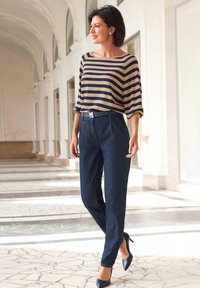 Gestreiftes beige und marineblaues Oberteil, kombiniert mit dunklen Denim-Hosen, die einen Gürtel mit einer glänzenden "M"-Schnalle und marineblauen High Heels auf einem marmorierten Boden tragen.