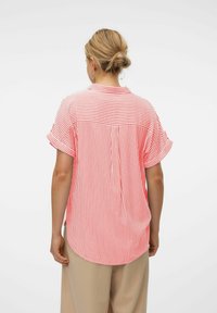 Camisa de manga corta a rayas rojas y blancas con detalle en el yugo trasero, fabricada con tela ligera. Dobladillo redondeado, corte relajado, sin cierres visibles.