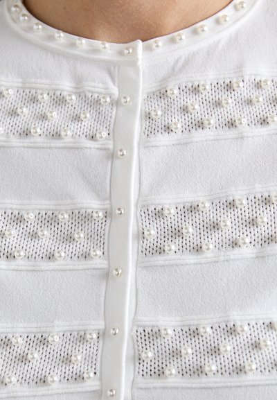Cardigan blanc avec des boutons en perles et des perles décoratives disposées en rayures maillées horizontales sur la poitrine et le col.