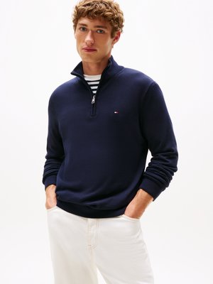 Marinefarbener Reißverschluss-Pullover aus weichem Stoff, mit einem hohen Kragen, gerippten Bündchen und einem kleinen Logodetail. Über einem gestreiften Shirt getragen.