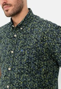 Button-up-Hemd mit dunklem Muster, das grüne, blaue und gelbe Sprenkel aufweist; mit einem spitzen Kragen und einem kleinen Logo.