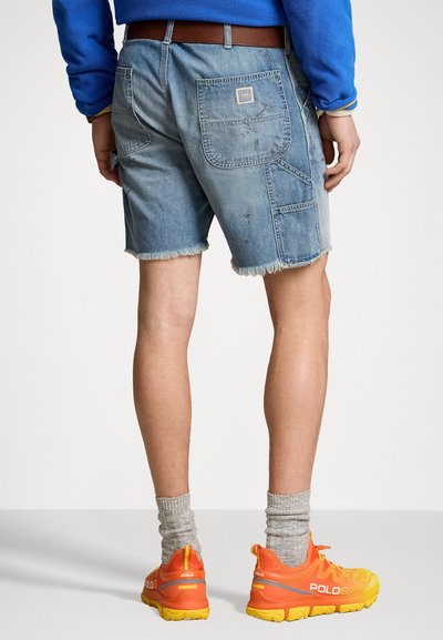 Pantalones cortos de mezclilla en azul claro con dobladillo deshilachado, que cuentan con múltiples bolsillos y un logotipo de parche, combinados con zapatillas deportivas de color naranja brillante y calcetines grises.