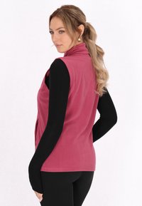 Gilet in pile rosa con colletto alto e senza maniche, abbinato a una maglietta nera a maniche lunghe. Presenta una texture liscia e un design aderente.