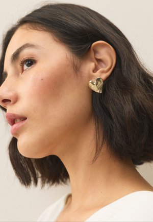 Goldenes herzförmiges Ohrstecker mit polierter Oberfläche, getragen am linken Ohr, zeigt eine glatte Textur und leichte Kurven.