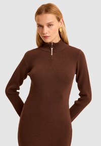 Abito marrone a costine con colletto alto e dettagli con mezza cerniera. Maniche lunghe e forma aderente, realizzato in un morbido materiale a maglia testurizzata.