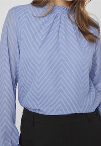 Blouse bleu clair à manches longues avec un motif en chevrons texturé et un col froncé, associée à un pantalon noir. Tissu lisse.