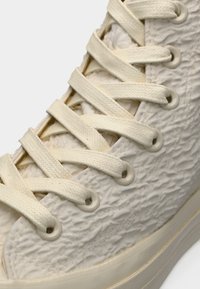 Υφασμένο κρεμ sneaker υψηλής κορυφής, με επίπεδα κρεμ κορδόνια, στρογγυλεμένη μύτη και λείο λαστιχένιο πάτο με διακριτικές λεπτομέρειες στην πάνω πλευρά.