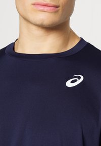 Camiseta deportiva de color azul marino con una textura suave, que presenta un cuello redondo y un acento de logo blanco en la zona de la clavícula.